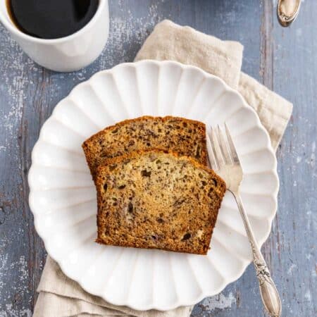 Two slice of banana bread on a white plate with a fork and a cup of coffee in the background.