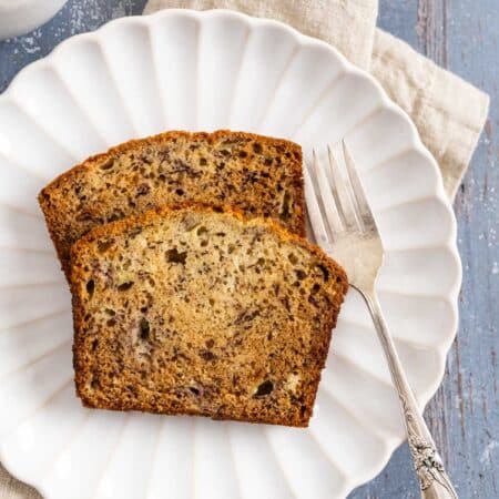 Two slice of banana bread on a white plate with a fork and a cup of coffee in the background.