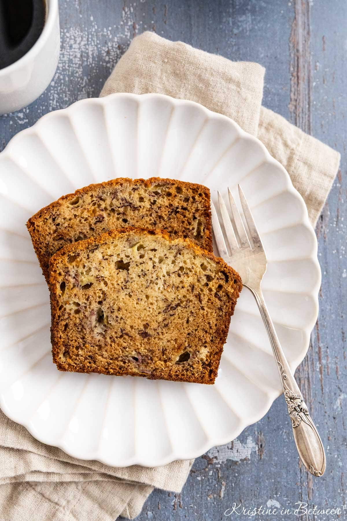 Two slice of banana bread on a white plate with a fork and a cup of coffee in the background.