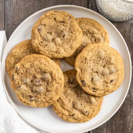 Chocolate chip cookies laying on a white plate with an eyelet napkin and a glass of milk in the background.