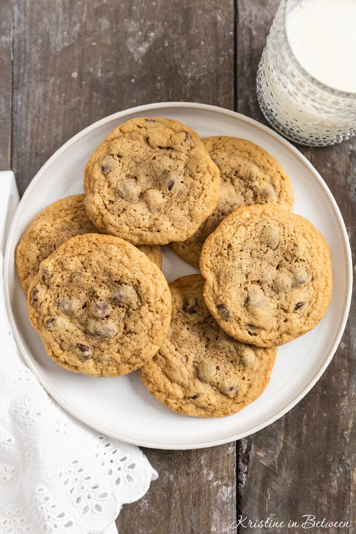 Chocolate chip cookies laying on a white plate with an eyelet napkin and a glass of milk in the background.