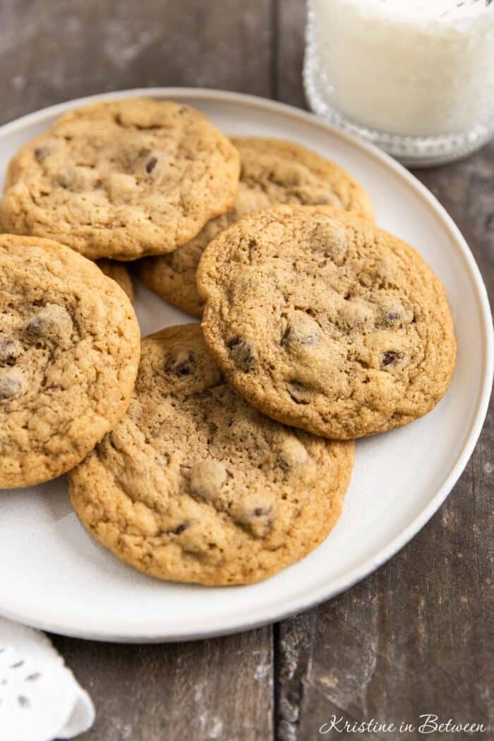 Chocolate chip cookies laying on a white plate with an eyelet napkin and a glass of milk in the background.