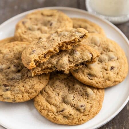 Chocolate chip cookies laying on a white plate with an eyelet napkin and a glass of milk in the background.