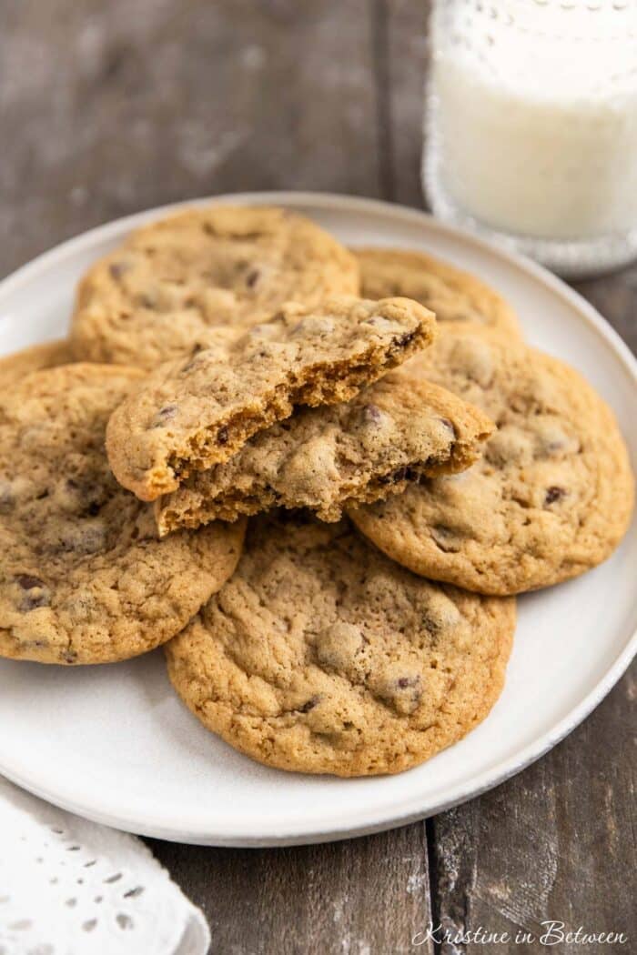 Chocolate chip cookies laying on a white plate with an eyelet napkin and a glass of milk in the background.