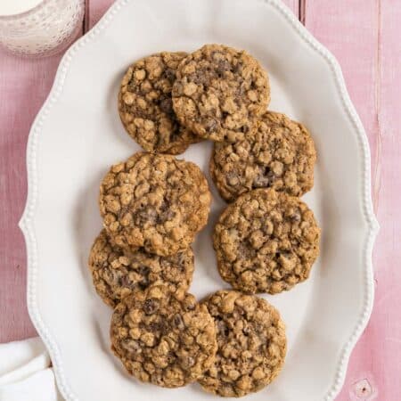 Oatmeal chocolate chip cookies stacked up on a white platter with a glass of milk in the background.