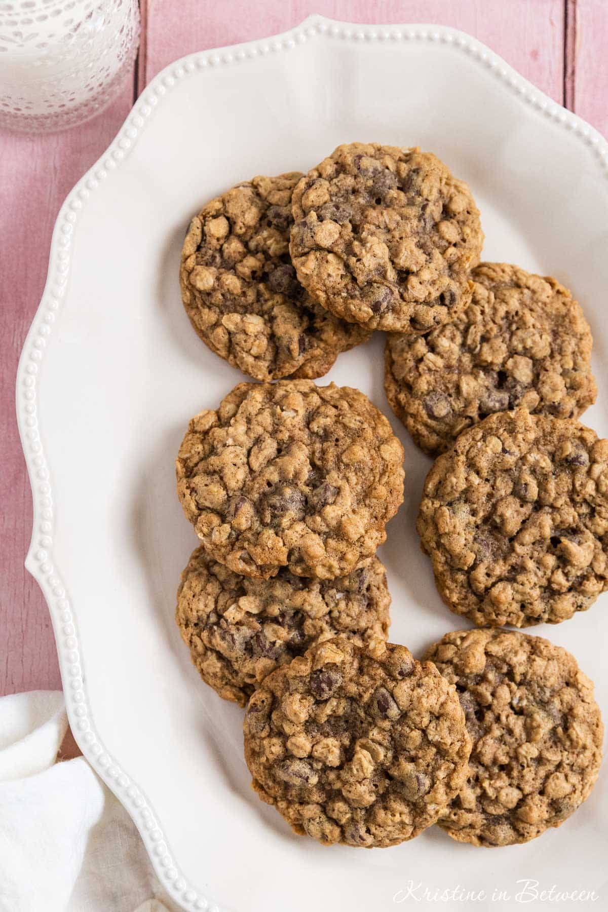 Oatmeal chocolate chip cookies stacked up on a white platter with a glass of milk in the background.