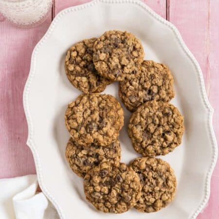 Oatmeal chocolate chip cookies stacked up on a white platter with a glass of milk in the background.