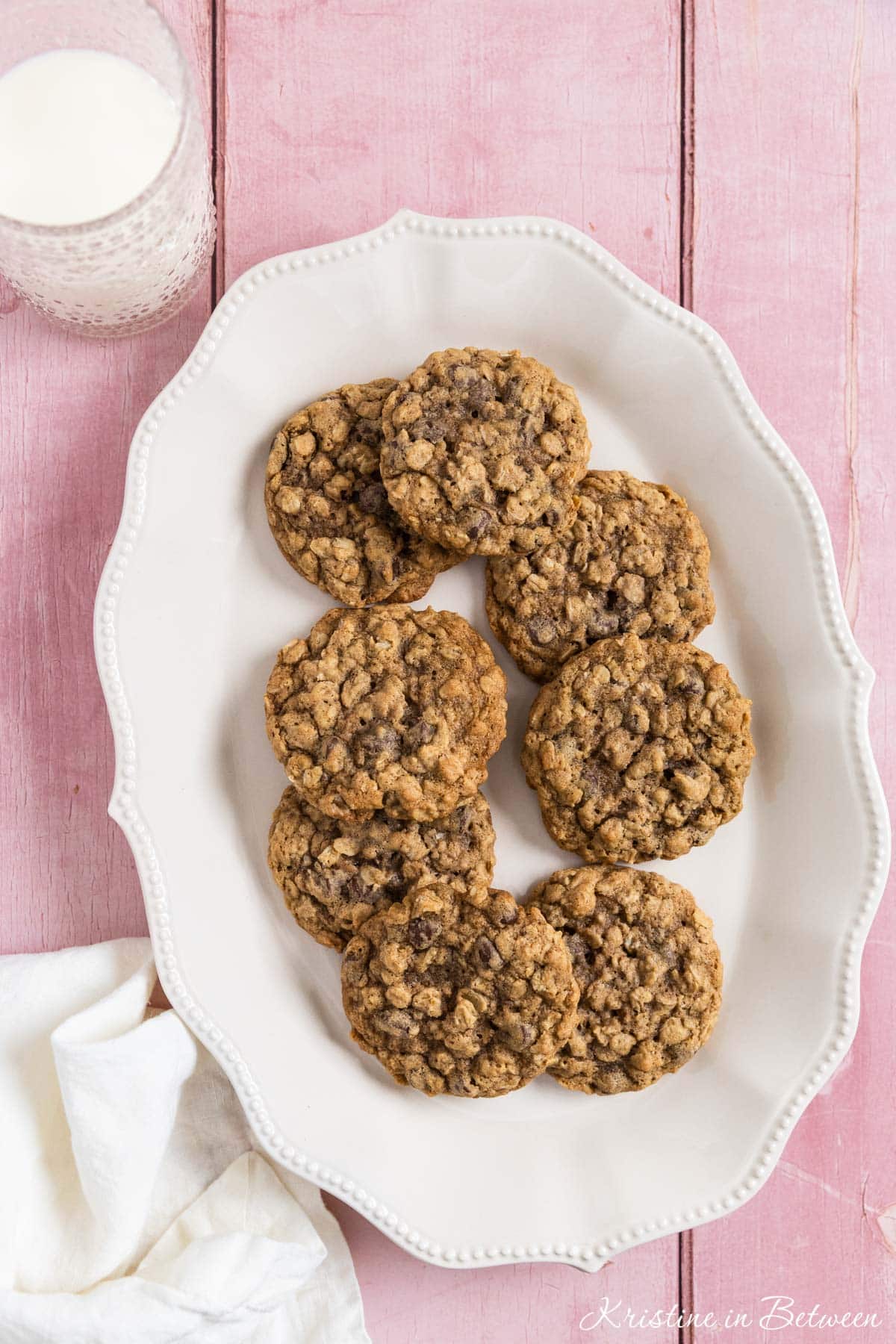 Oatmeal chocolate chip cookies stacked up on a white platter with a glass of milk in the background.