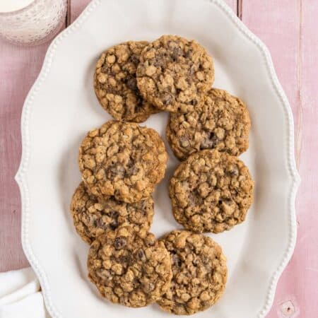 Oatmeal chocolate chip cookies stacked up on a white platter with a glass of milk in the background.