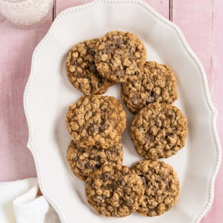 Oatmeal chocolate chip cookies stacked up on a white platter with a glass of milk in the background.