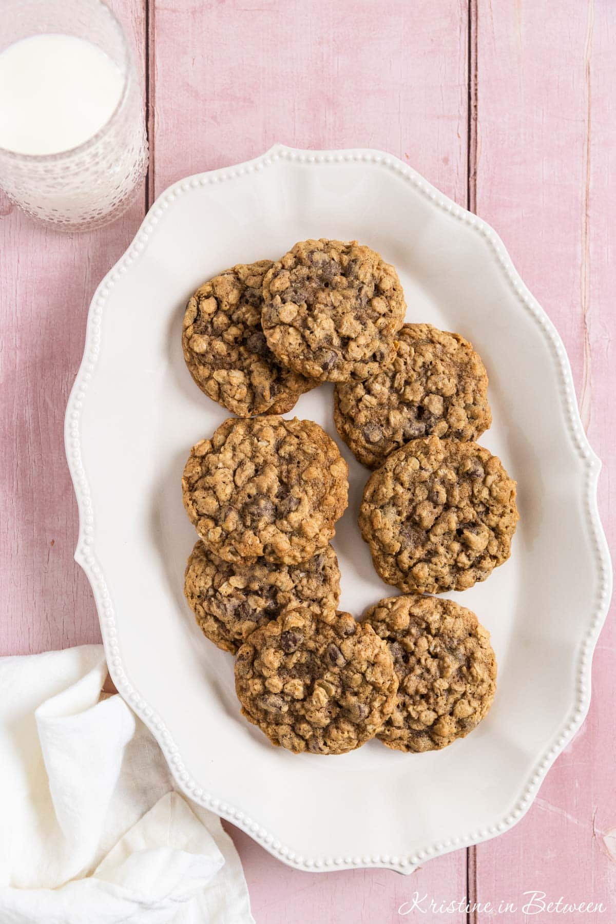 Oatmeal chocolate chip cookies stacked up on a white platter with a glass of milk in the background.