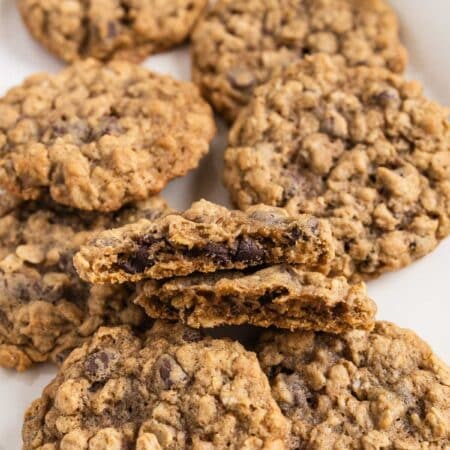 Oatmeal chocolate chip cookies stacked up on a white platter with a glass of milk in the background.