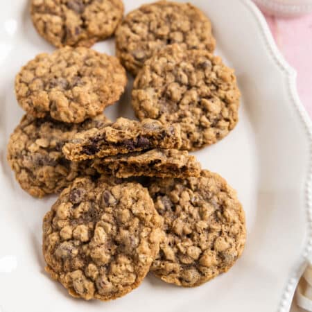 Oatmeal chocolate chip cookies stacked up on a white platter with a glass of milk in the background.