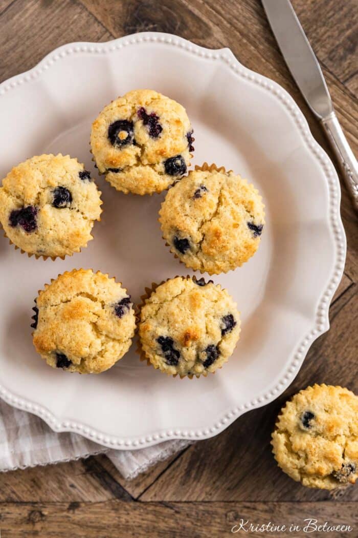 A plate of blueberry lemon muffins with one sitting on the side and a knife in the background.