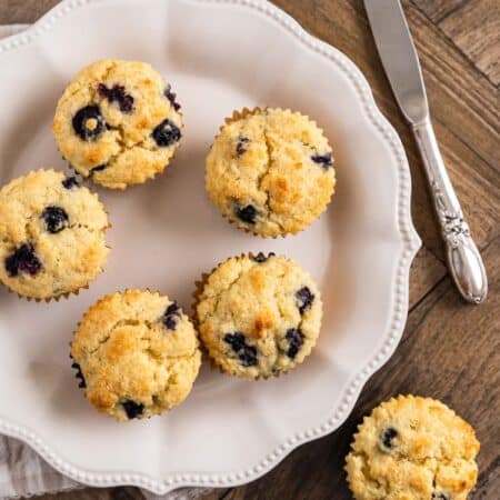 A plate of blueberry lemon muffins with one sitting on the side and a knife in the background.