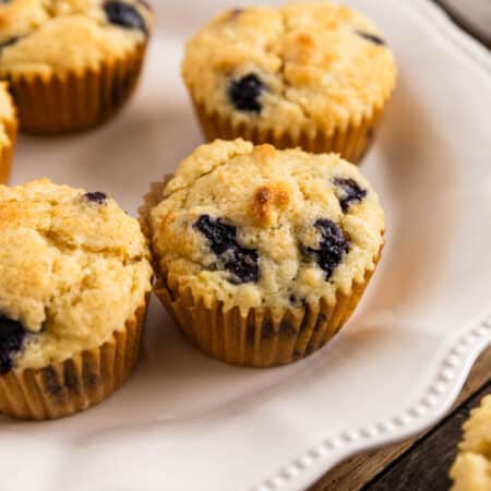 A plate of blueberry lemon muffins with one sitting on the side and a knife in the background.