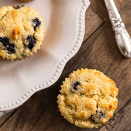 A plate of blueberry lemon muffins with one sitting on the side and a knife in the background.