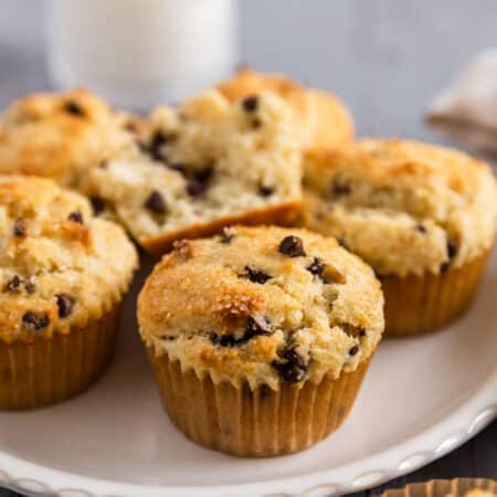 A chocolate chip muffin on a white plate with a glass of milk in the background.