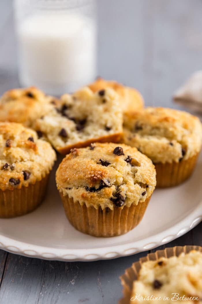 A chocolate chip muffin on a white plate with a glass of milk in the background.