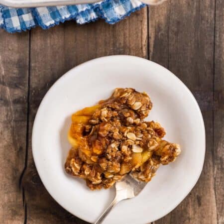 Peach crumble dished up on a white plate with a fork and the pan sitting behind it.