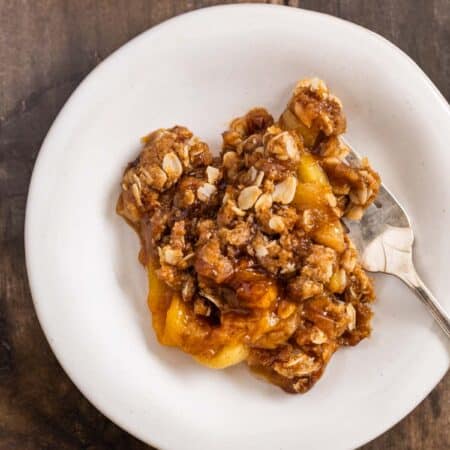 Peach crumble dished up on a white plate with a fork and the pan sitting behind it.