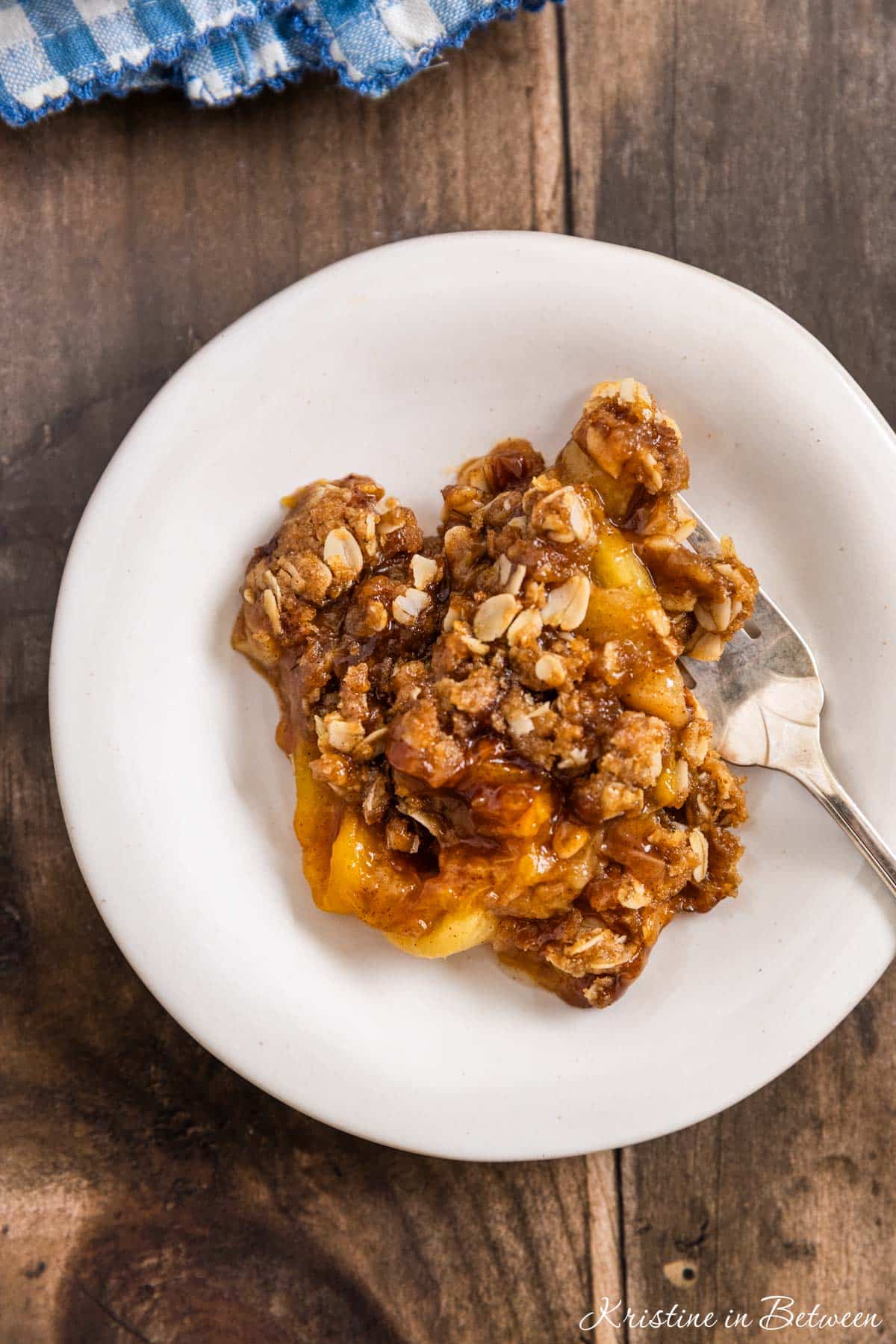 Peach crumble dished up on a white plate with a fork and the pan sitting behind it.