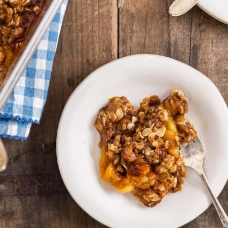 Peach crumble dished up on a white plate with a fork and the pan sitting behind it.