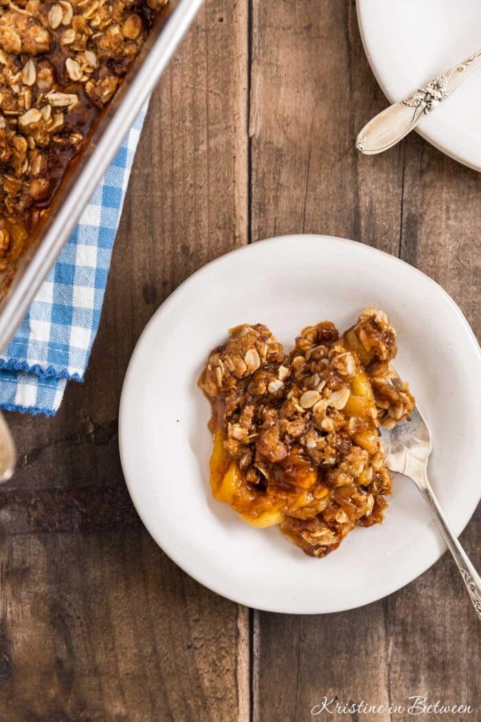 Peach crumble dished up on a white plate with a fork and the pan sitting behind it.