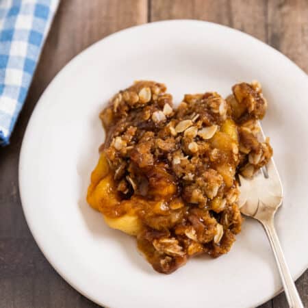 Peach crumble dished up on a white plate with a fork and the pan sitting behind it.