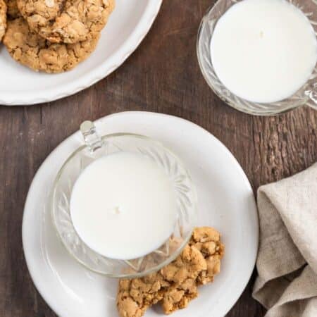 An oatmeal cooke broken in half on a white plate with a cup of milk next to it.