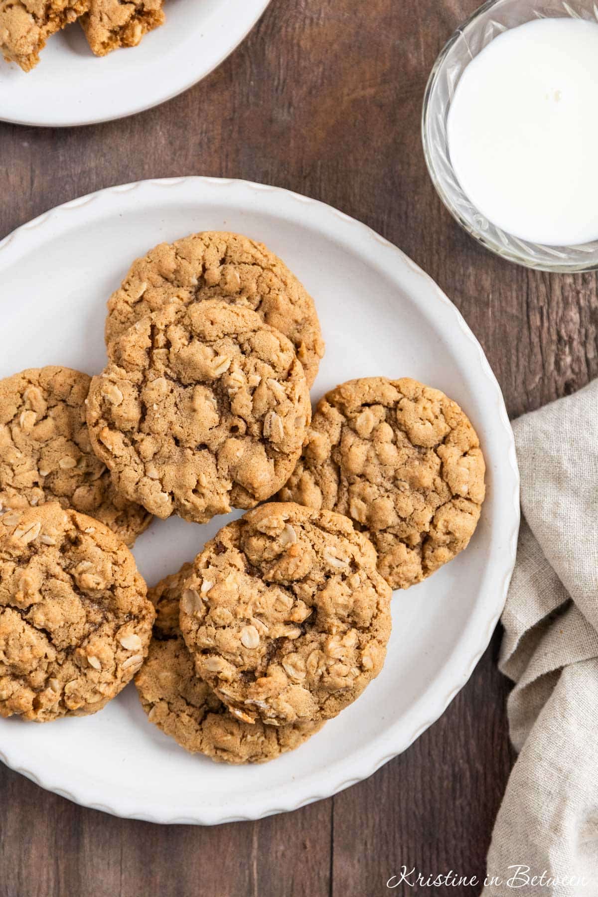 A plate of oatmeal cookies with a cup of milk and a tan napkin next to it.