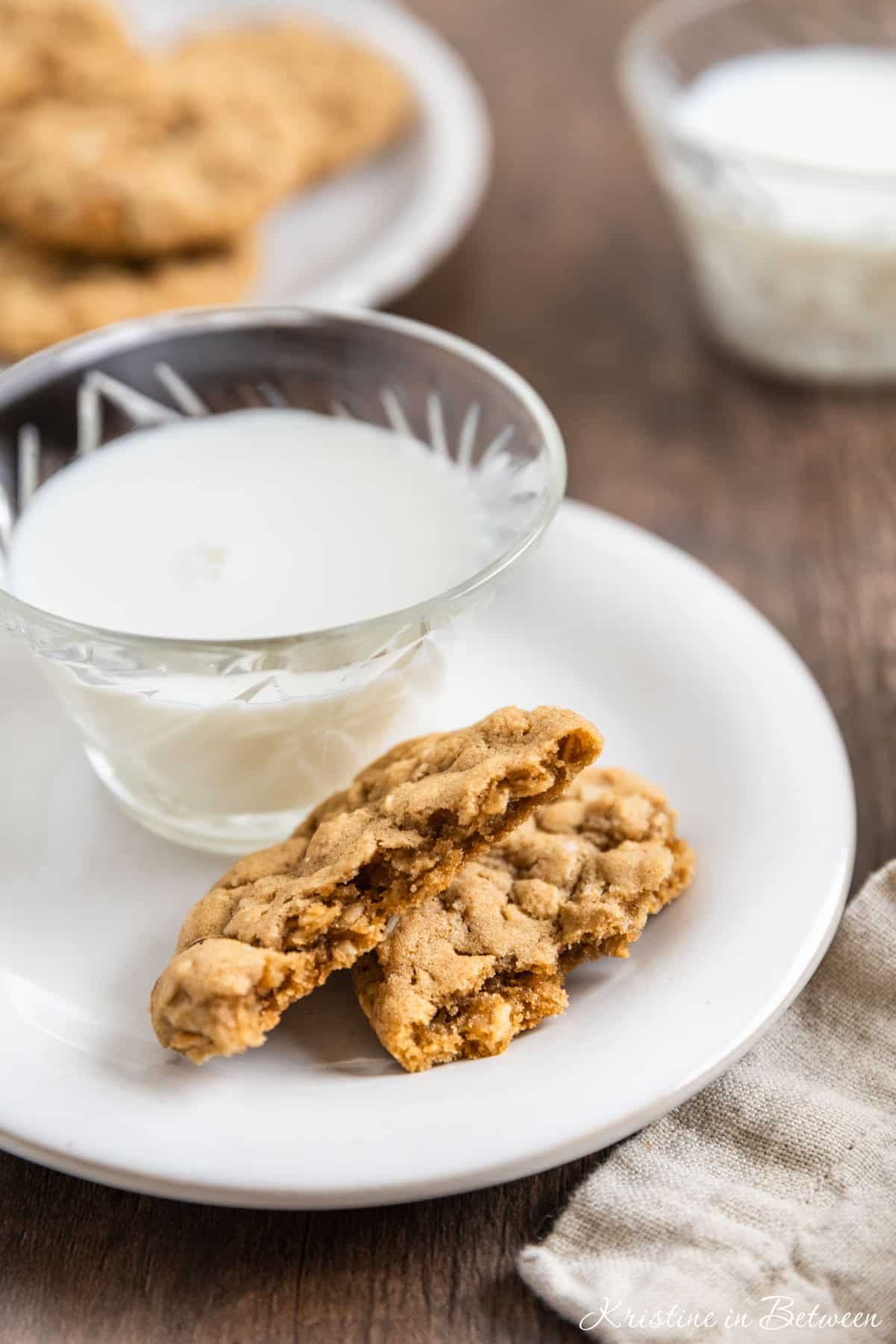 An oatmeal cooke broken in half on a white plate with a cup of milk next to it.