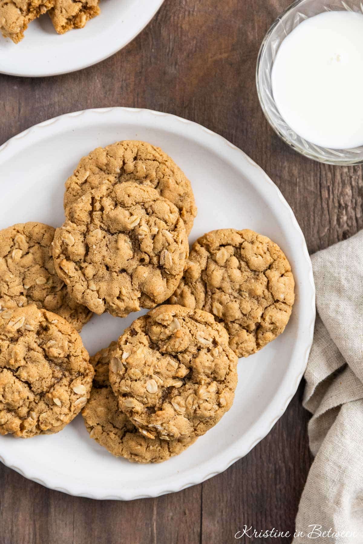 A plate of oatmeal cookies with a cup of milk and a tan napkin next to it.