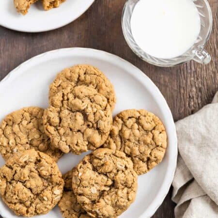 A plate of oatmeal cookies with a cup of milk and a tan napkin next to it.