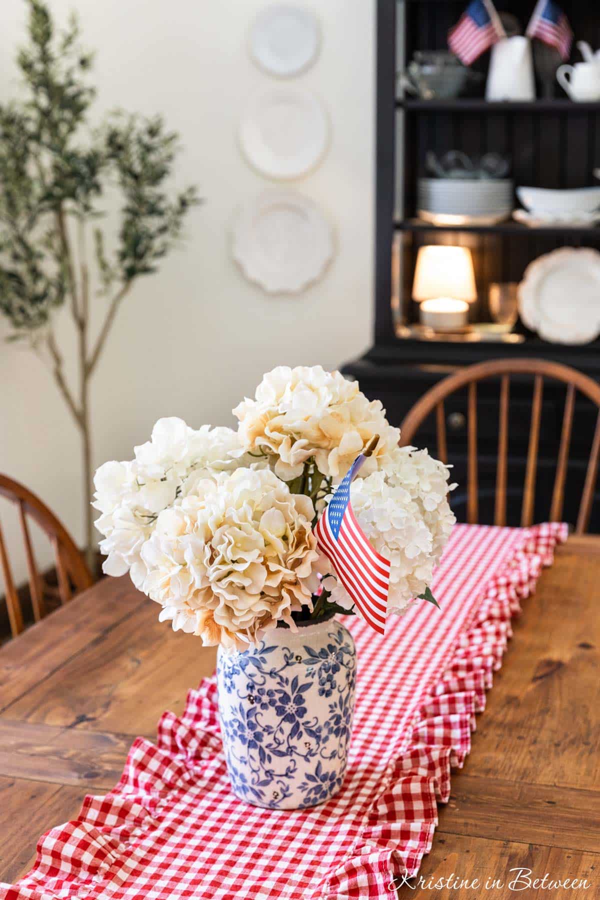 A vase of white flowers with a flag in them sitting on a table with a red gingham runner.