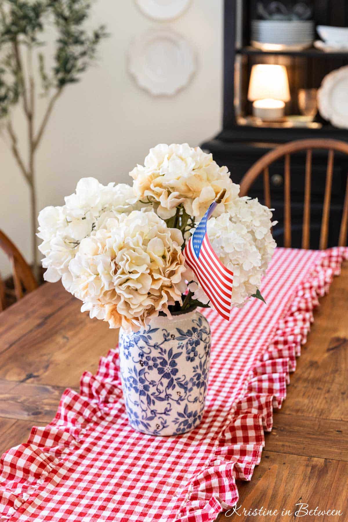 A vase of white flowers with a flag in them sitting on a table with a red gingham runner.