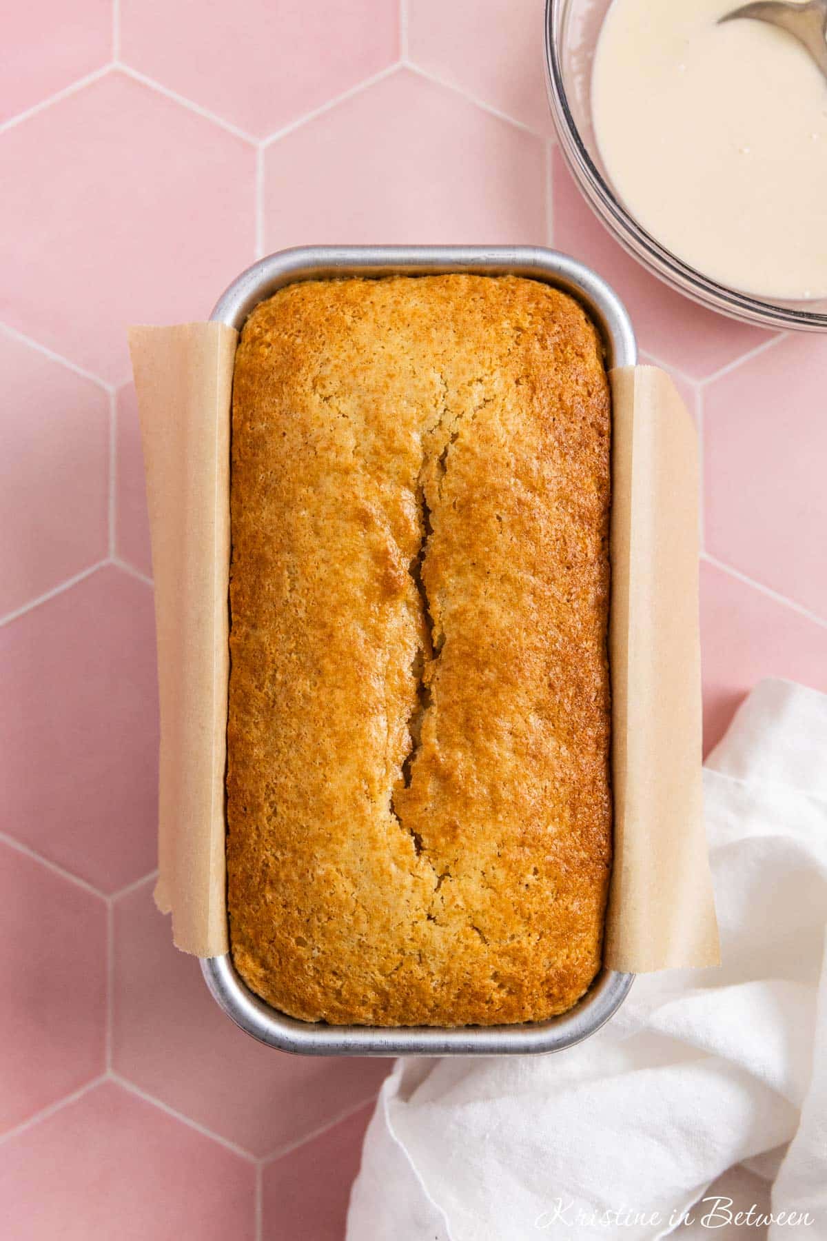 A glazed vanilla loaf in an aluminum bread pan with a white napkin next to it.