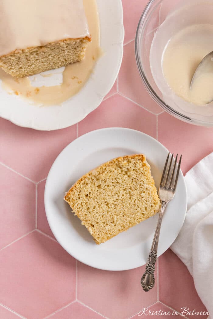 A slice of glazed vanilla loaf cake on a white plate with a fork next to it.
