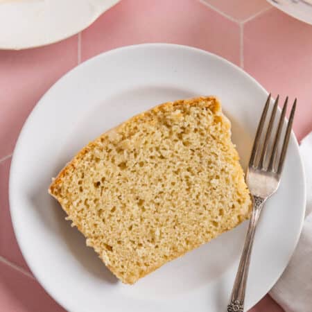 A slice of glazed vanilla loaf cake on a white plate with a fork next to it.