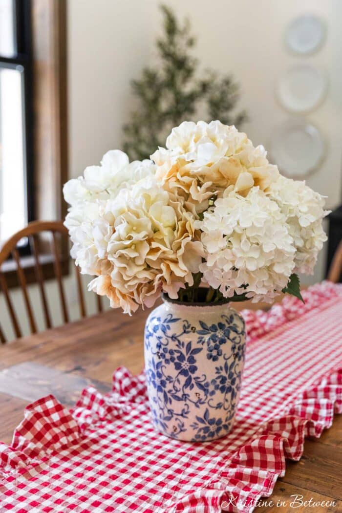 A vase of hydrangias on a wooden table with a red ghingam table runner.