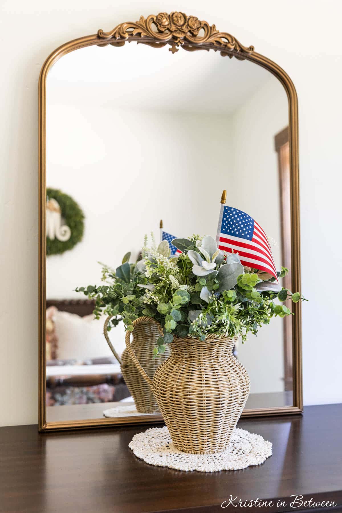 A woven vase of greenery with a flag in it in front of a gold trimmed mirror.