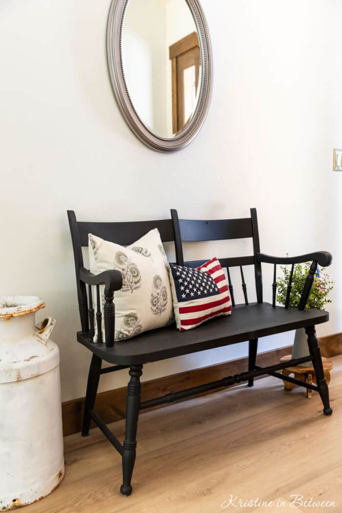 A black two-person bench sitting next to an old milk can with a flag pillow on it.