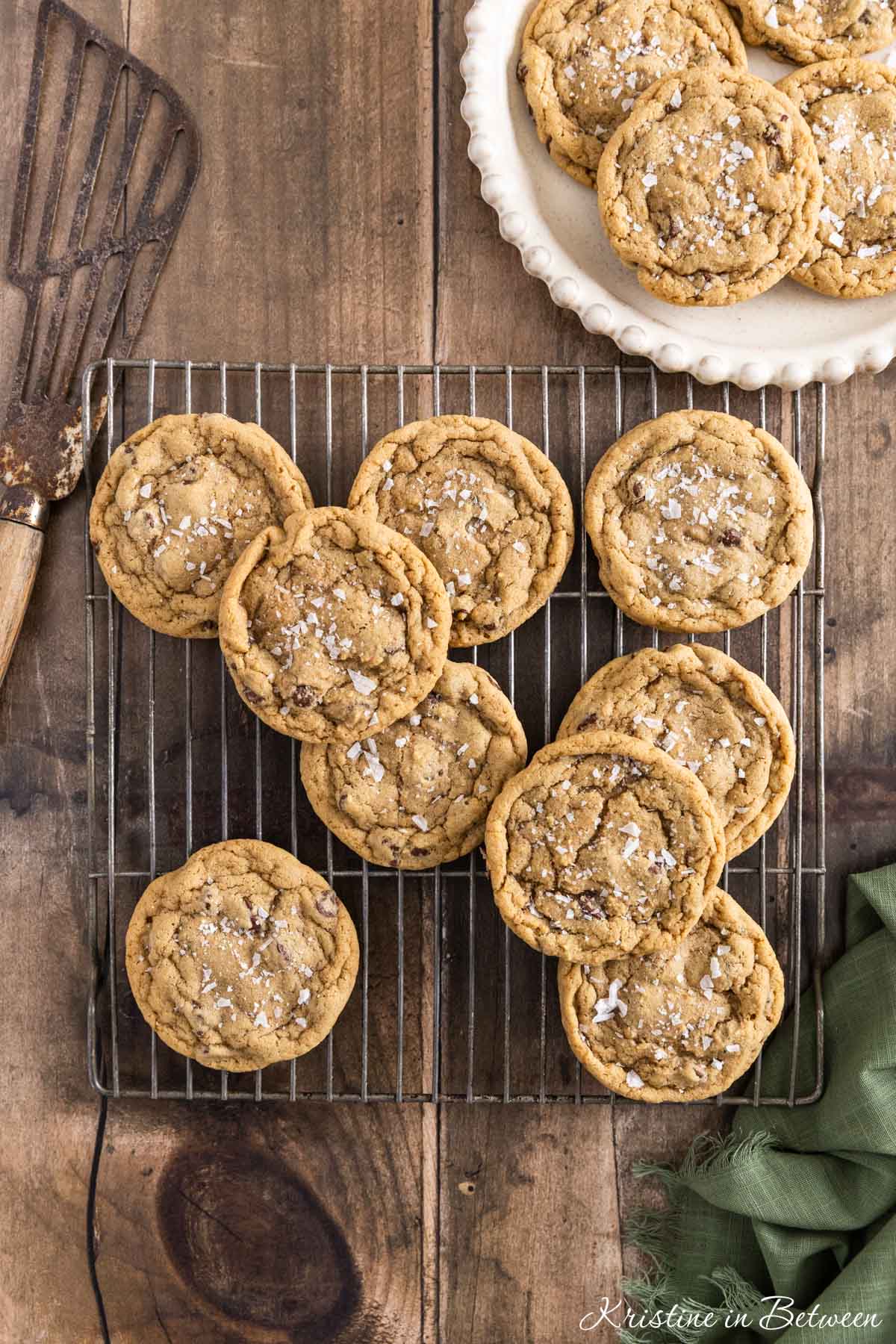 Chocolate chip cookies with sea salt sprinkled on top sitting on a wire rack with a green napkin next to them.