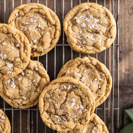 Chocolate chip cookies with sea salt sprinkled on top sitting on a wire rack with one broken in half to show the inside.