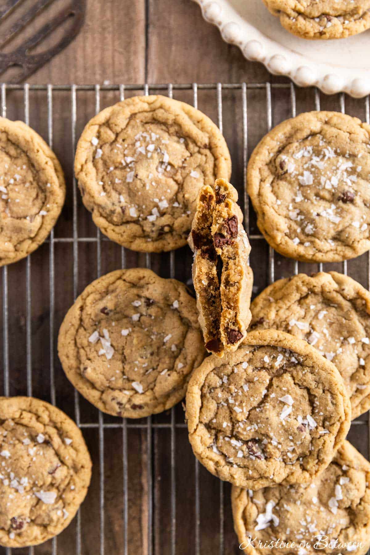 Chocolate chip cookies with sea salt sprinkled on top sitting on a wire rack with one broken in half to show the inside.