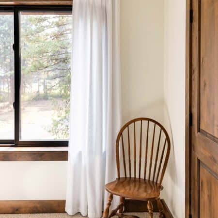 A room with white drapes hanging over a window with an antique chair in the corner.