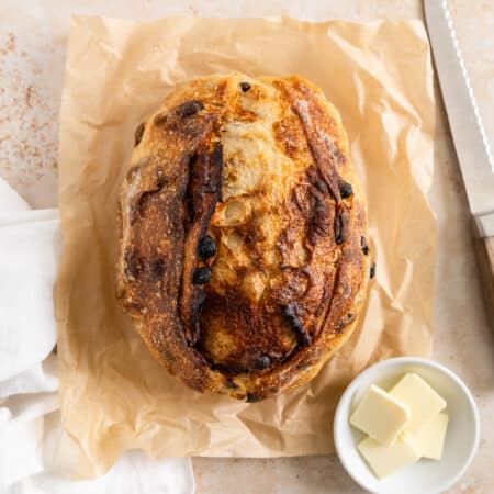 A loaf of cinnamon raisin sourdough bread sitting on a piece of parchment paper with butter next to it.