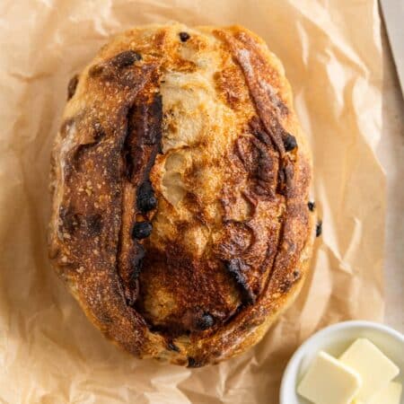 A loaf of cinnamon raisin sourdough bread sitting on a piece of parchment paper with butter next to it.