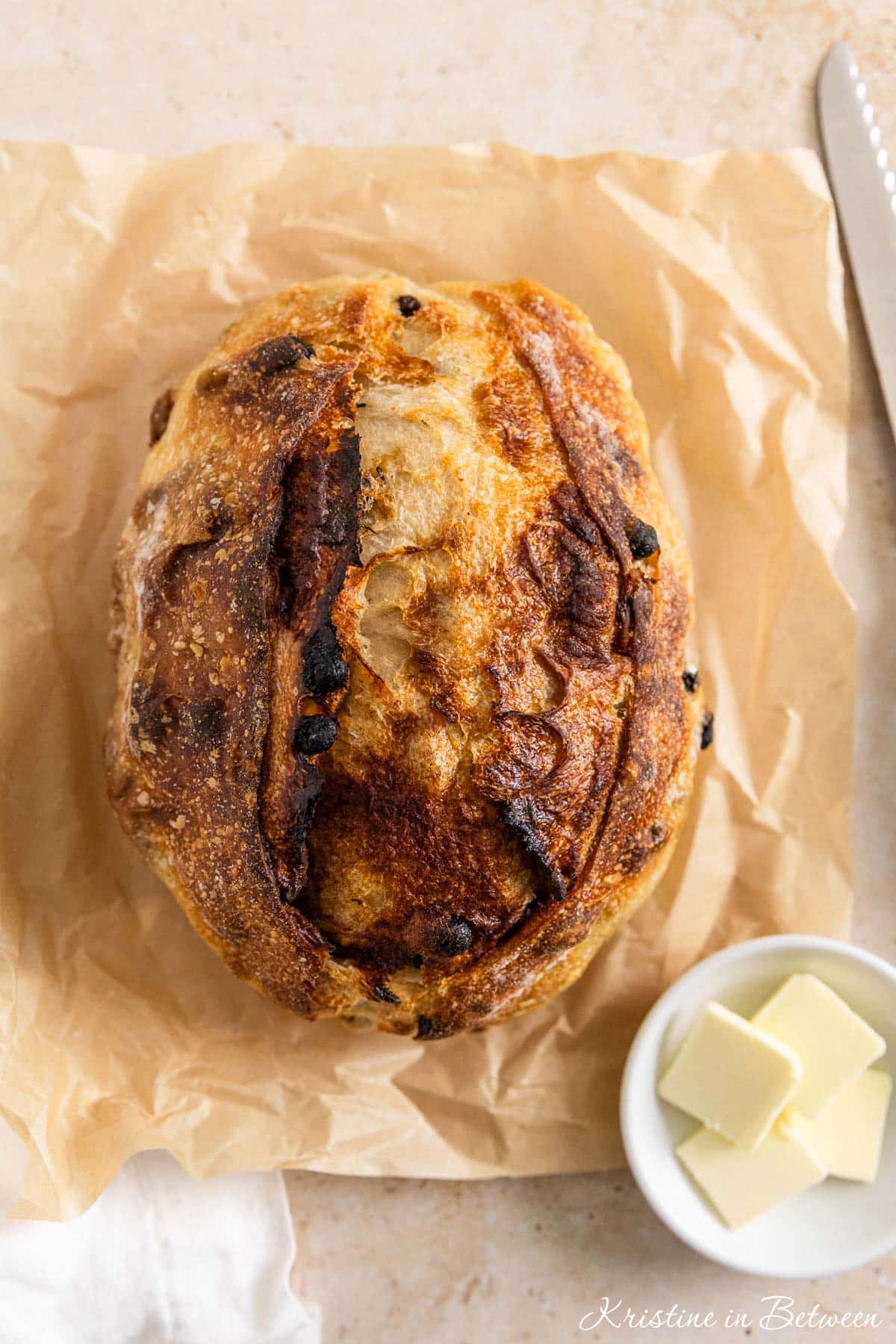 A loaf of cinnamon raisin sourdough bread sitting on a piece of parchment paper with butter next to it.