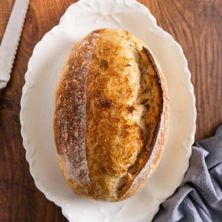 A whole loaf of sourdough bread on a white platter with a blue napkin sitting next to it.